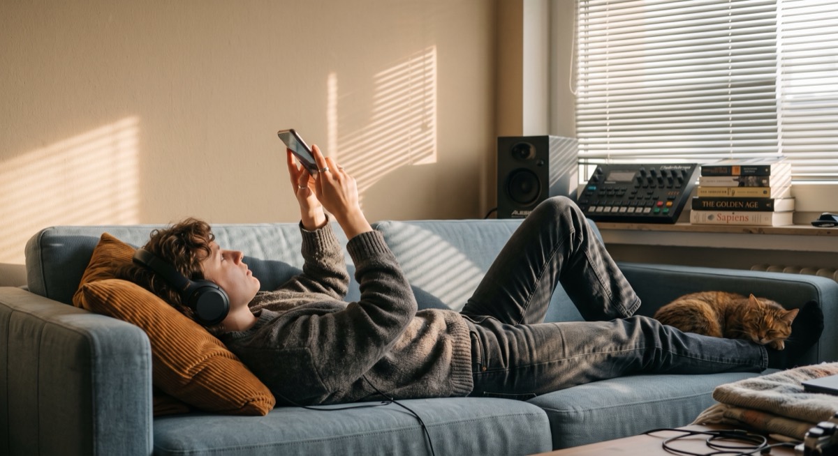 Someone lying along a blue sofa in headphones, holding a phone above their face, afternoon sun striping the wall through venetian blinds. A ginger cat sleeps on the armrest and a drum machine sits on the windowsill.