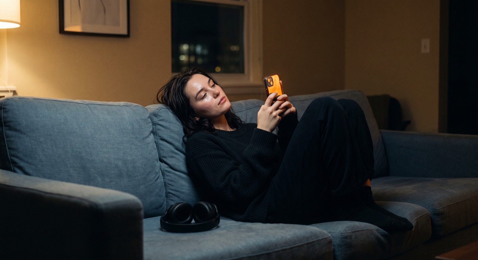 A woman lying back against the arm of a steel-blue sofa late at night, holding a phone up in one hand. The screen is the brightest thing in the room and throws warm amber across her face; a pair of headphones rests on the cushion beside her, and a lamp glows in the corner.